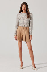 A woman is wearing a light-colored, textured, long-sleeved cropped jacket with a round neckline, decorated with buttons down the front. She pairs it with tan high-waisted pleated shorts. She completes the outfit with cream-colored high heels.